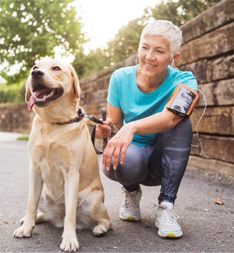 Senior woman with short hair and earphones kneels beside her happy dog during a walk on a sunny day.