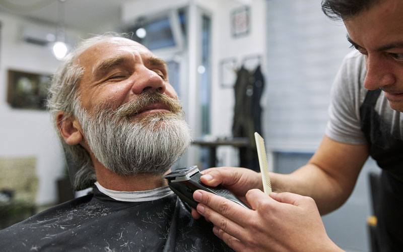 A barber trims a contented older mans beard in a modern barbershop.