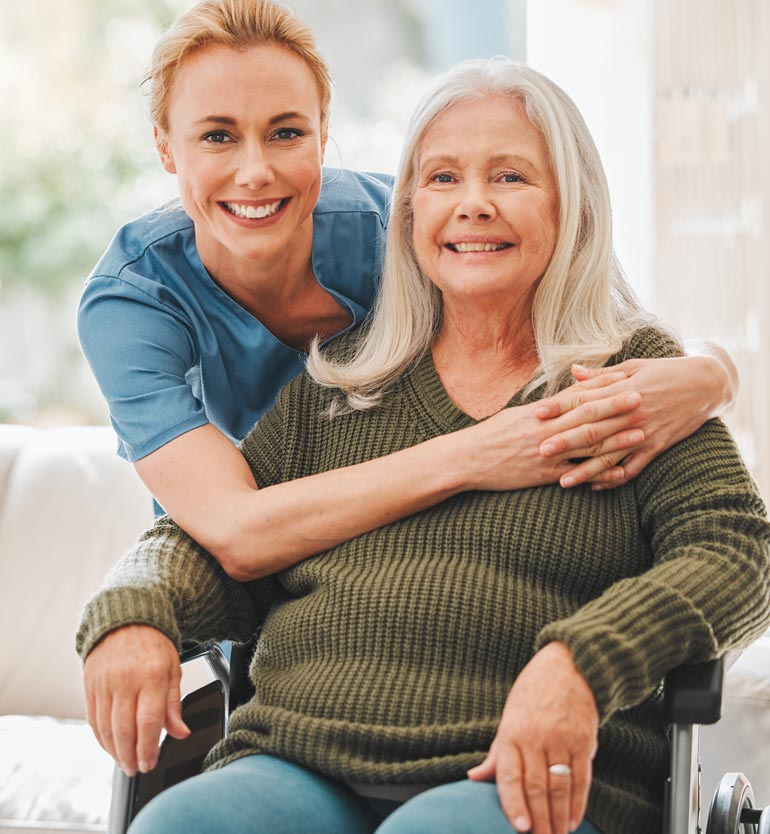 A smiling caregiver embraces a senior woman in a wheelchair inside a cozy home setting.