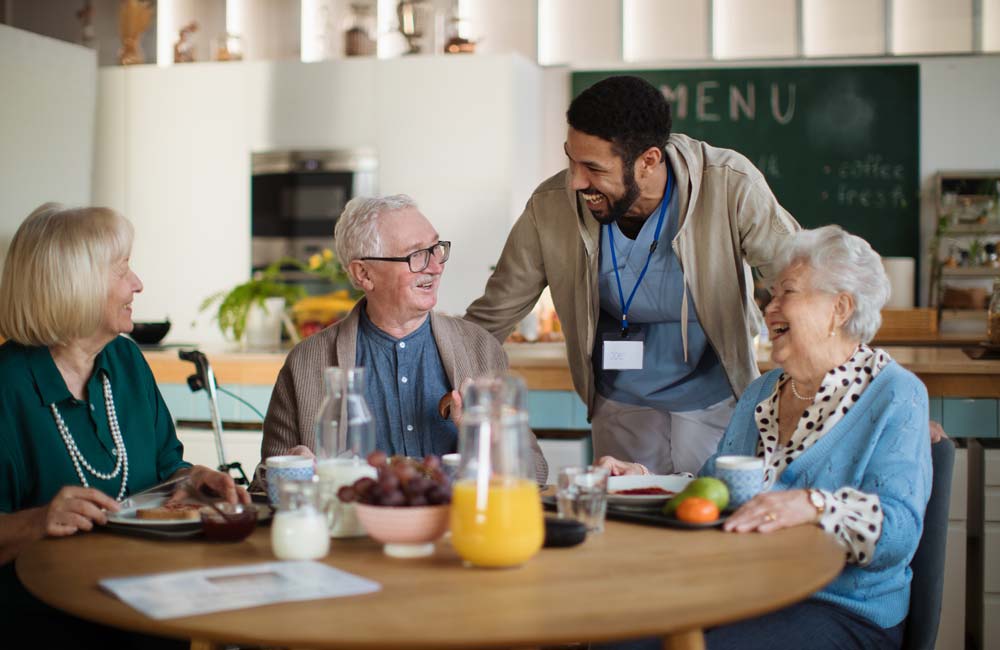 Caregiver engaging in conversation with three senior individuals at a dining table in a care home.