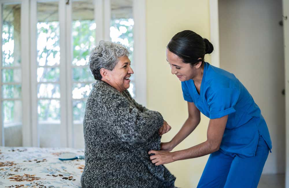 Caregiver in blue scrubs helps smiling elderly woman put on a cardigan near a window.