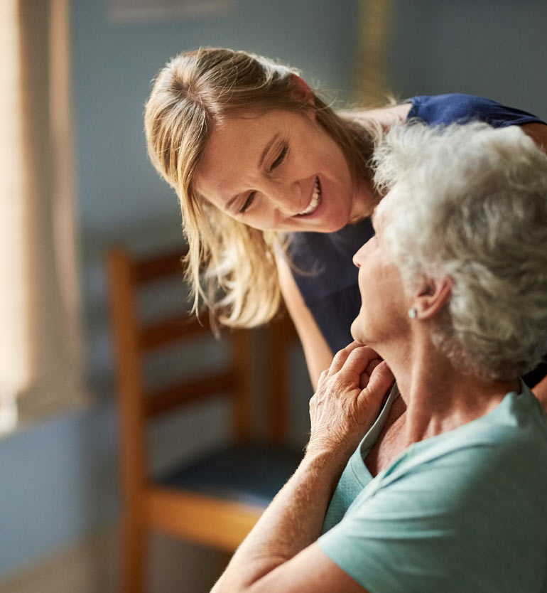 A caregiver smiling warmly at a seated elderly woman in a well-lit room.