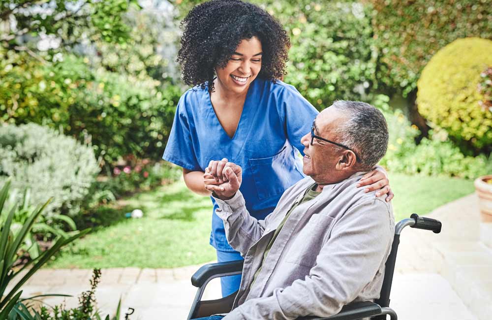 Caregiver in blue scrubs smiling and interacting with senior man in wheelchair outdoors.