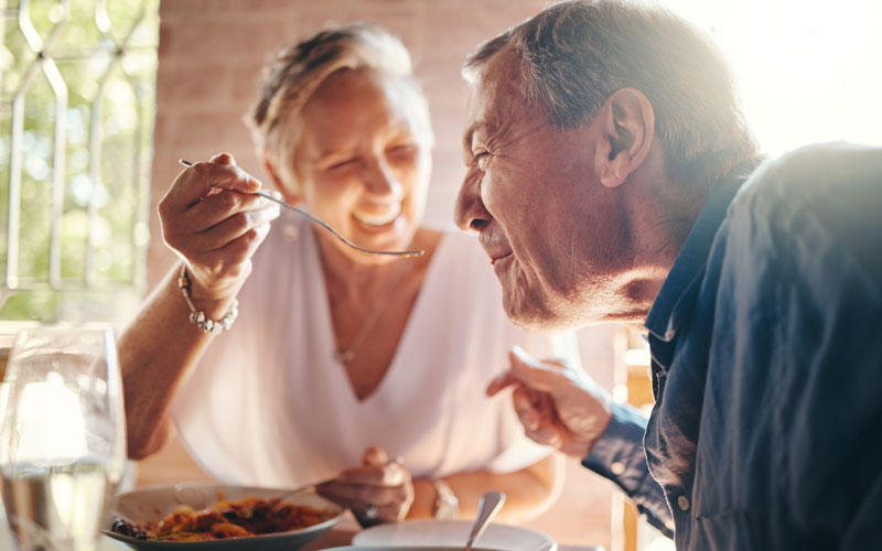 Elderly couple enjoying a meal together, with the woman feeding the man while both smile warmly.