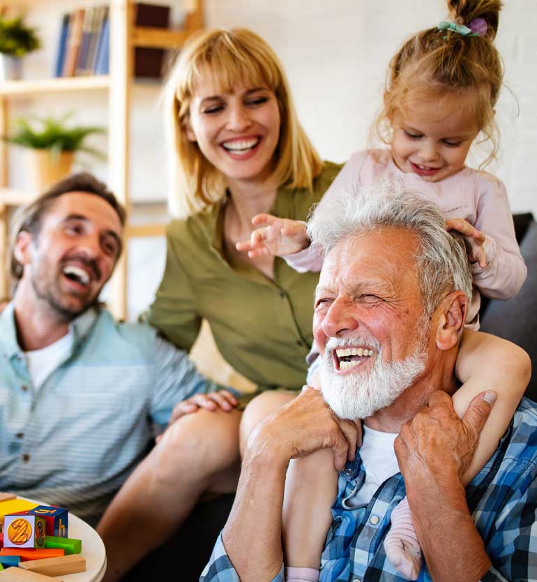 Elderly man laughing with young child on shoulders, surrounded by smiling family members.
