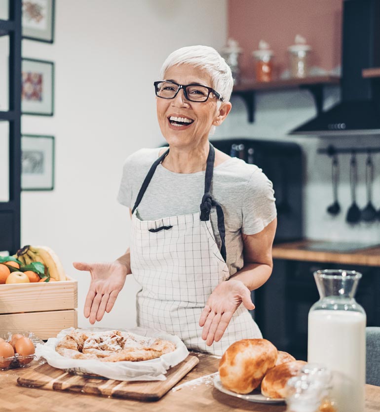 A smiling elderly woman in an apron proudly displays her freshly baked goods in a cozy kitchen.