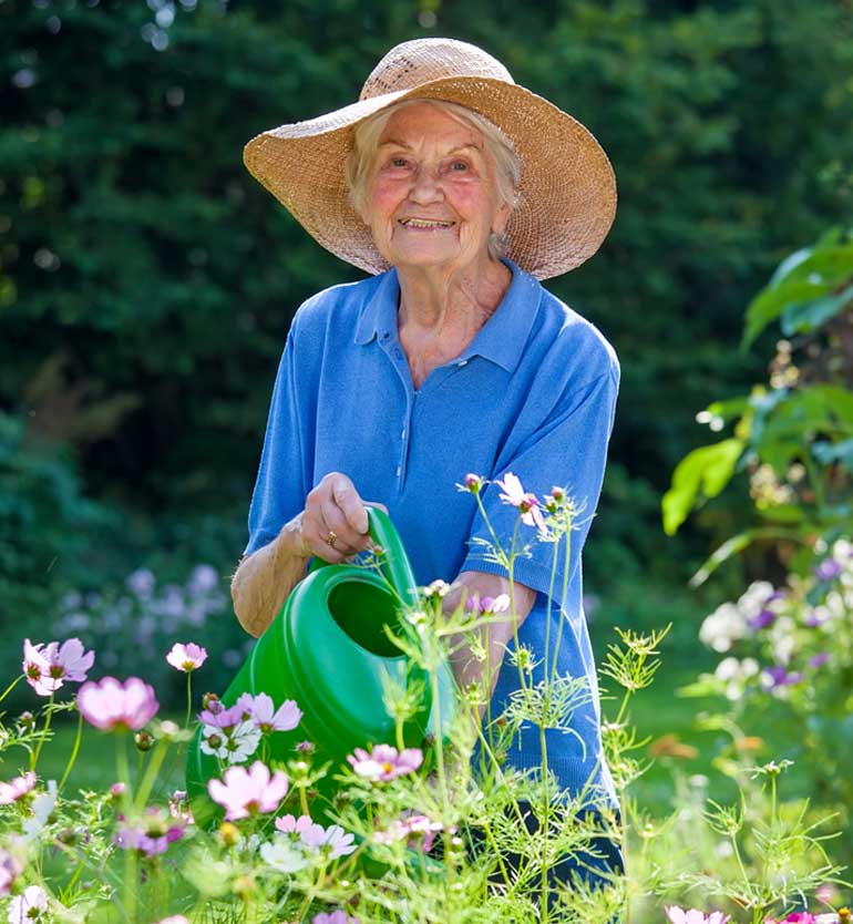 Elderly woman in a straw hat and blue shirt smiling while watering flowers in a bright garden.