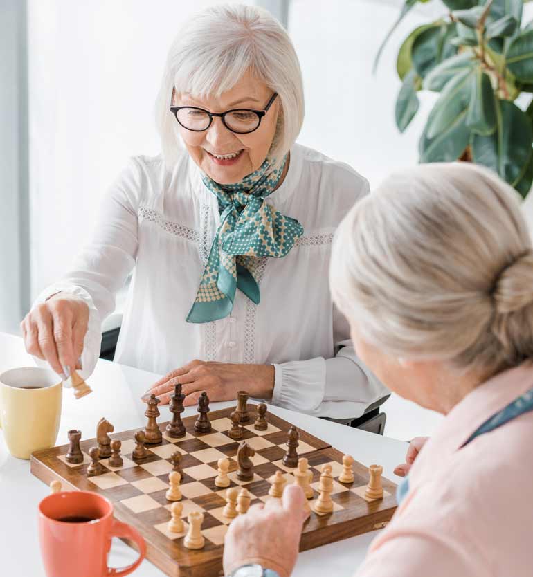 Two elderly women enjoying a game of chess at a bright, indoor setting, with coffee cups nearby.