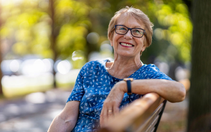Happy senior woman with glasses smiling while sitting on a park bench on a sunny day.