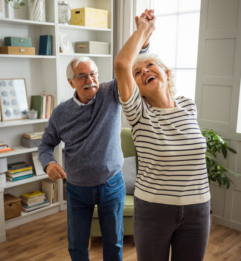 Senior couple dancing joyfully in their living room with bookshelves in the background.