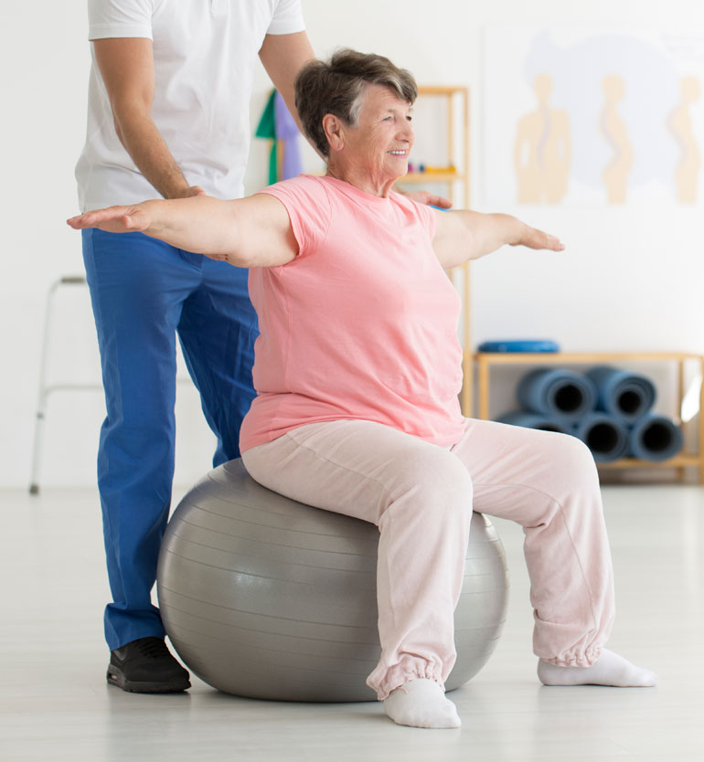 A senior woman in a pink shirt doing physical therapy exercises on a stability ball with a therapist.