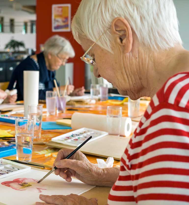 A senior woman concentrates on painting a flower at a colorful art class for older adults.