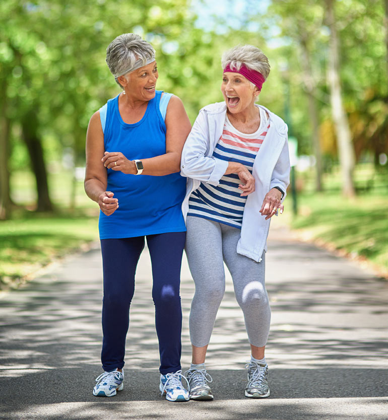 Two senior women laughing and exercising together on a park path on a sunny day.