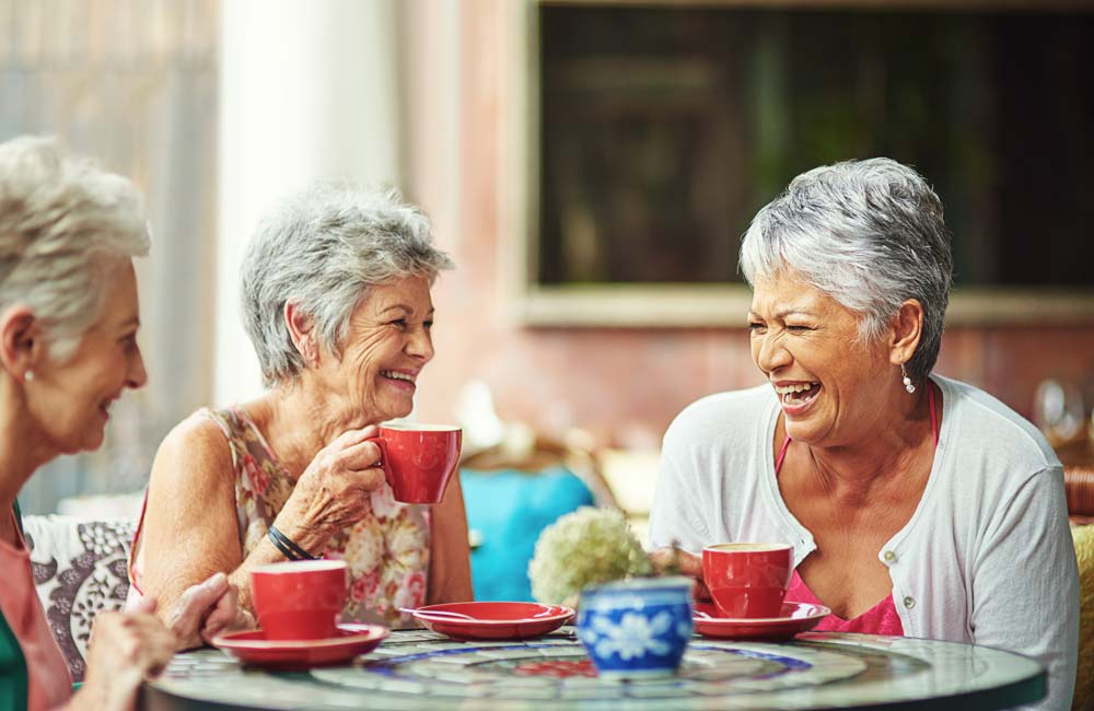 Three senior women enjoying coffee together, laughing and smiling at a table with red cups.