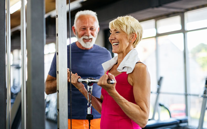 Two smiling seniors exercising with gym equipment in a bright, modern fitness center.